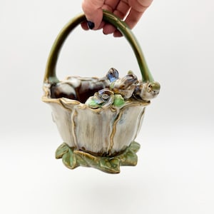 Pode incluir: Uma cesta de cerâmica com alça e base verdes, com um design floral. A cesta é principalmente bege com detalhes marrons e verdes, e pequenas figuras de pássaros estão pousadas na borda. A cesta é segurada por uma pessoa.