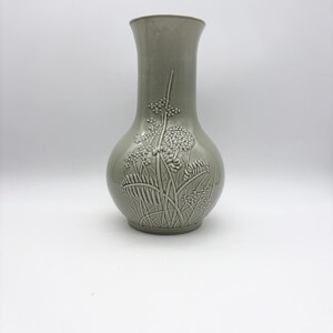 May include: A sage green ceramic vase with a bulbous body and flared neck. The vase features a raised relief design of stylized plants and flowers. The vase is a decorative home decor item.
