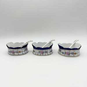 以下が含まれることがあります： 青と金の縁取りと花柄の白い磁器の小さなボウルが3つ。各ボウルには小さな白いスプーンが付いています。
