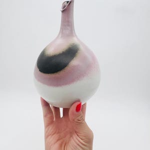 Może przedstawiać: Ręcznie wykonany ceramiczny wazon o pękatym korpusie i smukłej szyi. Wazon ma gradient kolorów, przechodzący od bieli u podstawy do różu, a następnie ciemnoszarego wiru. Wazon jest trzymany przez dłoń.