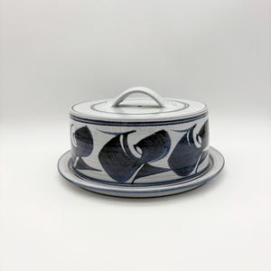 May include: Ceramic cake stand with lid. The stand and lid are white with a blue and black abstract design. The lid has a handle for easy lifting. The stand sits on a matching plate.