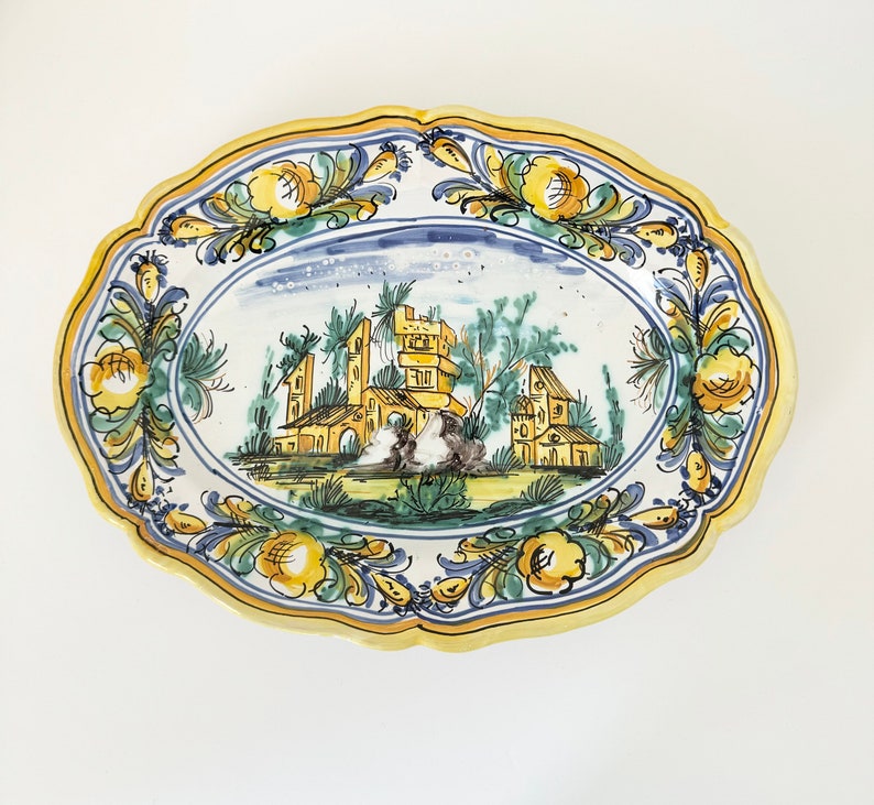 Pu&ograve; includere: Un piatto da portata ovale in ceramica con un bordo floreale giallo, blu e verde. Il centro del piatto presenta una scena dipinta a mano di un piccolo villaggio con edifici e alberi.