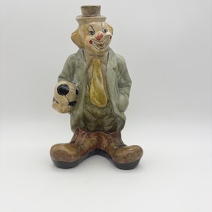 Könnte beinhalten: Keramik-Clownfigur, bekleidet mit einem grünen Mantel, einer gelben Krawatte und braunen Stiefeln, hält einen schwarz-weißen Fußball. Der Clown hat ein bemaltes Gesicht mit roter Nase und einem kleinen Hut. Die Figur steht auf einer weißen Oberfläche.