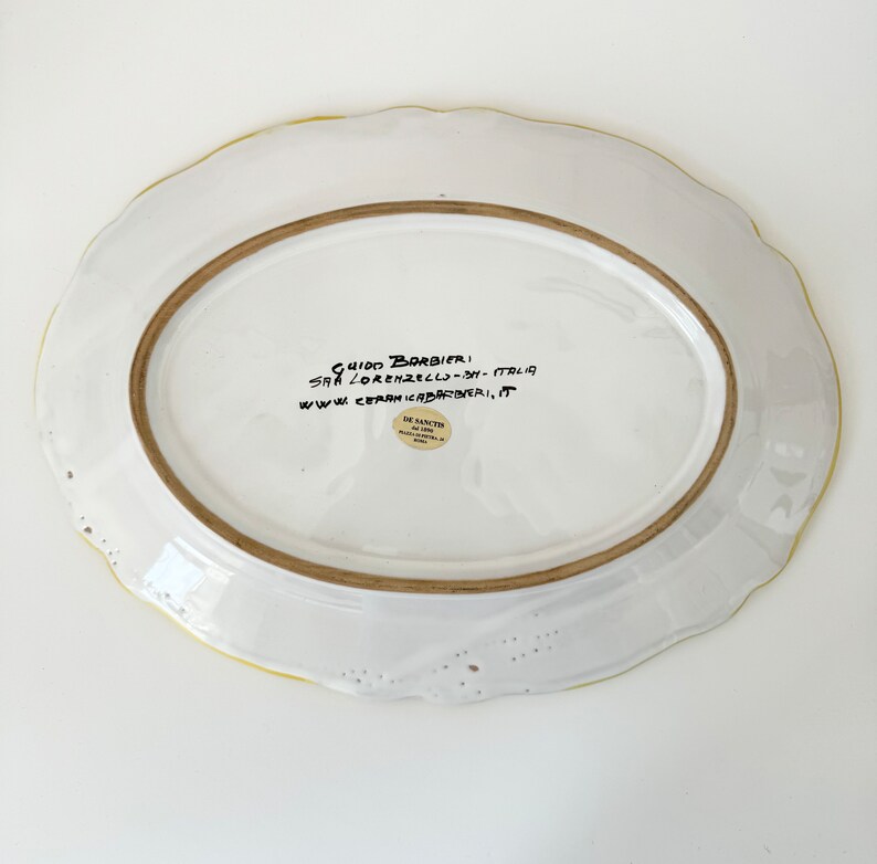 Pu&ograve; includere: Piatto da portata ovale in ceramica bianca con bordo giallo e un'iscrizione manoscritta sul fondo: "GUIDO BARBIERI, SAN LORENZELLO - OM - ITALIA, WWW.CERAMICABARBIERI, IT". &Egrave; visibile anche un piccolo adesivo rotondo con il testo "DE SANCHIS".