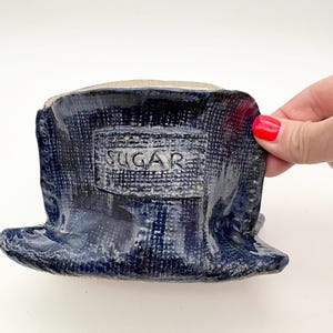 Puede incluir: Un recipiente de cerámica esmaltada en azul oscuro con una superficie texturizada similar a la tela. Una placa rectangular en la parte frontal presenta la palabra SUGAR en letras mayúsculas. El recipiente tiene una base ancha y un borde.