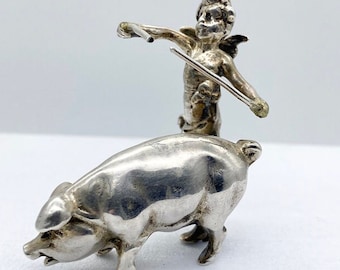 May include: A silver figurine of a cherub playing a violin on top of a pig.