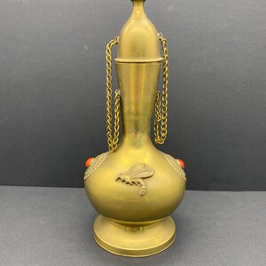 Op de afbeelding: Een gouden metalen fles met een decoratief ontwerp van een libel en rode accenten. De fles heeft een ketting die aan de dop is bevestigd.