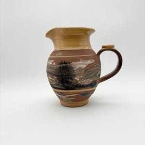 May include: A handcrafted ceramic pitcher with a landscape design. The pitcher features a brown base, a tan rim, and a dark brown handle. The landscape design includes a tree, clouds, and various colors. The pitcher is a decorative item.