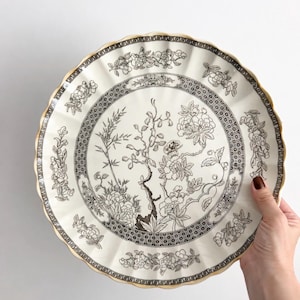 May include: A round ceramic plate with a scalloped edge and a gold-toned rim. The plate features a black and white botanical illustration of flowers, bamboo, and a butterfly, surrounded by a geometric border pattern on a cream-coloured background.