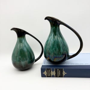 May include: Two ceramic pitchers with a glossy, teal and dark green glaze. The pitchers have a teardrop shape with a curved handle and a dark brown rim. One pitcher is smaller than the other. The larger pitcher rests on a blue book.