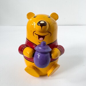 May include: A yellow bear figurine with a red shirt, holding a small purple pot. The bear has a smiling expression with a black nose and mouth, and is painted with a glossy finish. The figure is seated with its legs extended forward.