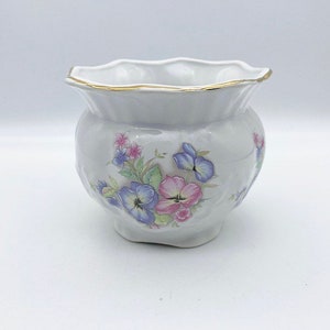May include: A white ceramic planter with a scalloped rim and a gold trim. The planter features a floral design with pink, blue, and green flowers.