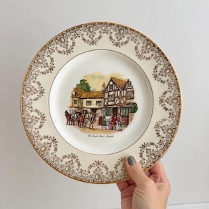 May include: A round ceramic plate with an off-white base and a gold filigree border. The centre features a colourful illustration of a timber-framed building with horses and people, labelled with the text Old Coach House - Bristol in black script.