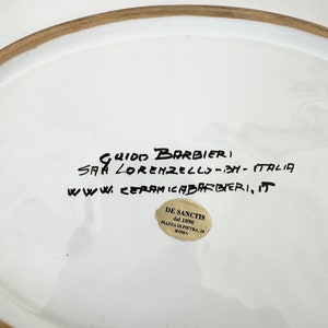 Pu&ograve; includere: Piatto in ceramica bianca con bordo marrone e una scritta a mano in inchiostro nero che recita "Guido Barbieri, S.A.A. Lorenzello - BH - Italia, www.ceramicabarbieri, it". Un adesivo dorato con testo nero recita "De Sanctis dal 1890 Piazza di Pietra, 24 Roma".