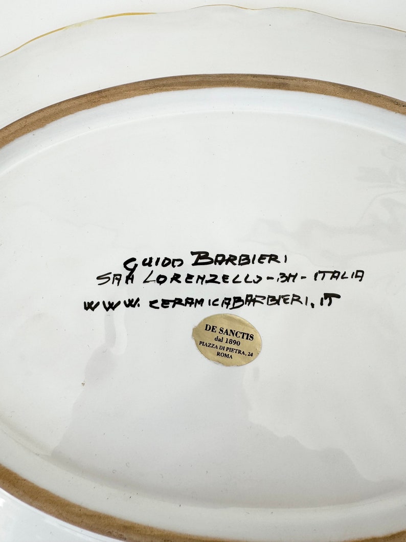 Pu&ograve; includere: Piatto in ceramica bianca con bordo marrone e una scritta a mano in inchiostro nero che recita "Guido Barbieri, S.A.A. Lorenzello - BH - Italia, www.ceramicabarbieri, it". Un adesivo dorato con testo nero recita "De Sanctis dal 1890 Piazza di Pietra, 24 Roma".