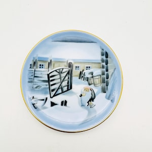 Könnte beinhalten: Ein kleiner, runder, blau-weißer Porzellanteller mit goldfarbenem Rand. Der Teller zeigt eine Winterszene mit einem kleinen, weißen Haus, einem Holzzaun und einer Figur in roter Mütze, die im Schnee geht.