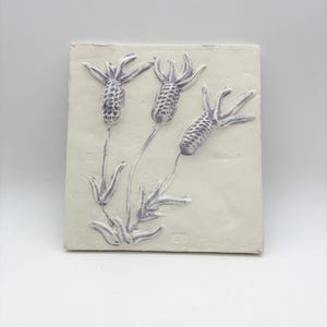 Könnte beinhalten: Quadratische, cremefarbene Keramikfliese mit einem erhabenen Reliefdesign von drei Lavendelblütenstängeln. Die Blüten und Stiele sind in einem gedämpften Lila gehalten und weisen detaillierte Texturen auf. Die Fliese ist ein dekoratives Stück.