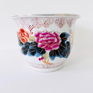 Op de afbeelding: Een witte keramische bloempot met een uitlopende rand, versierd met een levendig bloemmotief. De bloemen zijn in roze, rood en geel, met donkerblauwe bladeren. De rand heeft een decoratief patroon en een gouden rand.