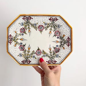 May include: An octagonal ceramic serving platter with a cream-coloured base, featuring a hand-painted floral pattern of pink and blue flowers, green leaves, and small orange blossoms. The design includes dark red bows and a speckled black dot border.