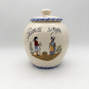 May include: Cream-colored ceramic jar with lid, featuring a blue floral pattern on the rim and lid. The jar has a hand-painted scene of two figures and the words "Poubelle de table". A decorative kitchen storage container.
