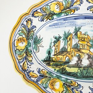 Pu&ograve; includere: Un grande piatto da portata ovale in ceramica con un design dipinto a mano. Il piatto presenta un bordo floreale blu e giallo con una scena centrale di un edificio giallo con un tetto blu e alberi verdi. Il piatto &egrave; perfetto per servire pasta, insalate o altri piatti.