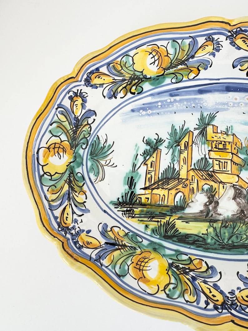 Pu&ograve; includere: Un grande piatto da portata ovale in ceramica con un design dipinto a mano. Il piatto presenta un bordo floreale blu e giallo con una scena centrale di un edificio giallo con un tetto blu e alberi verdi. Il piatto &egrave; perfetto per servire pasta, insalate o altri piatti.