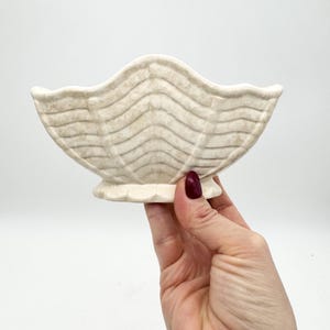 Puede incluir: Un cuenco de cerámica blanco con un diseño ondulado y acanalado. El cuenco tiene forma de concha y tiene una base pequeña.