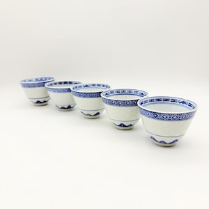 May include: Five white porcelain teacups with blue geometric patterns. The cups have a flared shape and a delicate blue border design. The cups are arranged in a row, showcasing their intricate details and classic aesthetic.