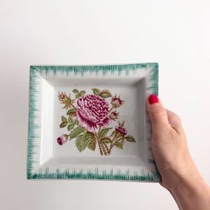 May include: A rectangular white ceramic tray featuring a central illustration of a blooming pink rose with green leaves and thorny stems. The rim of the tray is decorated with a textured green border pattern that mimics a fringe or grass-like design.