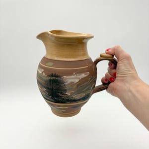 May include: A ceramic pitcher featuring a tan glazed rim and a brown body decorated with a painted landscape scene of a tree and swirling clouds. The pitcher has a curved handle and a wide pouring spout, showcasing a hand-painted artistic design.