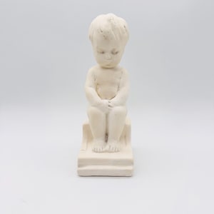 Può includere: Piccola statua color crema di un bambino seduto con le mani giunte in grembo. Il bambino è seduto su una piccola piattaforma a gradini. La statua ha una finitura opaca ed è ambientata su uno sfondo bianco.