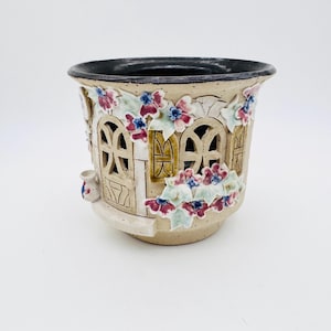 May include: A beige ceramic planter shaped like a house, featuring arched windows and floral embellishments. The planter has a dark rim and a small, decorative pot on the side. The flowers are red, blue, and white.