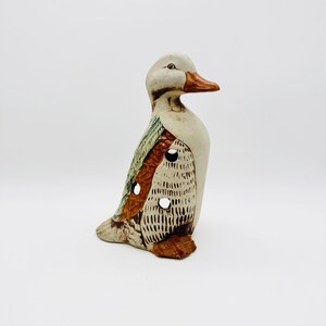 May include: A ceramic duck figurine with brown, green and white paint. The duck has a textured surface and several holes.
