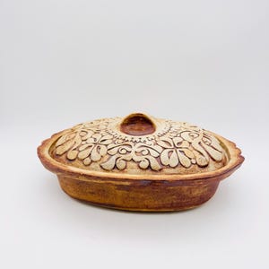 Puede incluir: Una fuente ovalada de cerámica con tapa. La tapa y el borde presentan un relieve floral. La fuente es principalmente marrón con detalles crema. La tapa tiene un pequeño asa marrón.