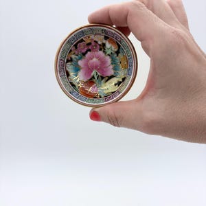 May include: A small, round, decorative dish with a floral design. The dish features a pink peony in the center, surrounded by other flowers and gold accents. The rim has a geometric pattern and a gold border.