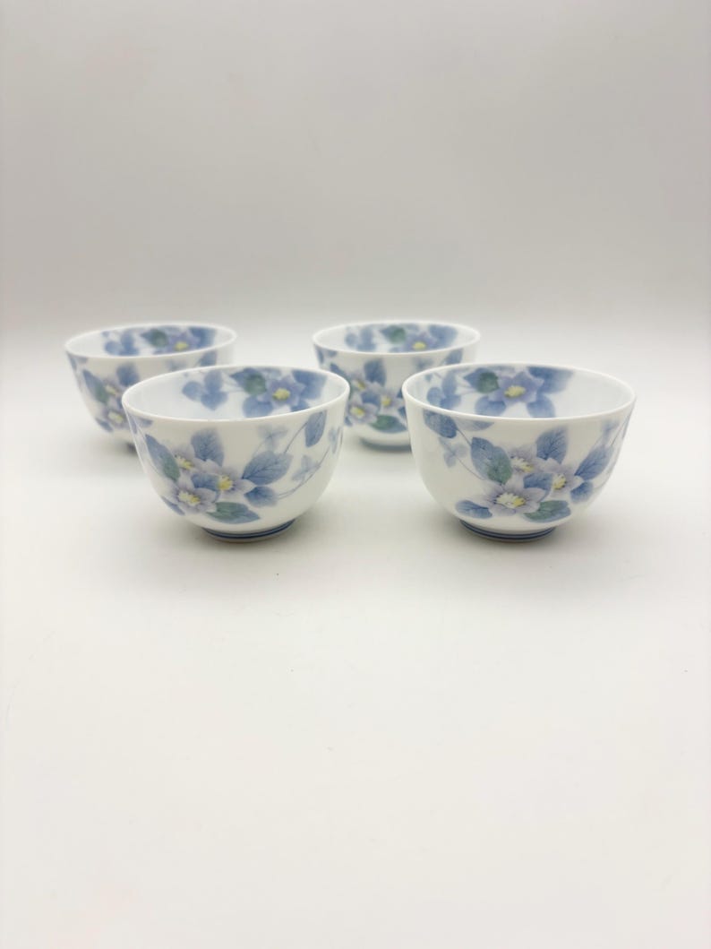 K&ouml;nnte beinhalten: Vier wei&szlig;e Keramikschalen mit einem Blumendesign mit blauen und gelben Blumen und gr&uuml;nen Bl&auml;ttern. Die Schalen sind auf wei&szlig;em Hintergrund angeordnet. Das Blumenmuster wiederholt sich auf jeder Schale.