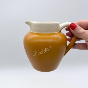 May include: A small, mustard-yellow ceramic pitcher with a cream-colored rim and handle. The word "Cheddar" is written in cursive on the front of the pitcher. The pitcher is designed for serving sauces or liquids.