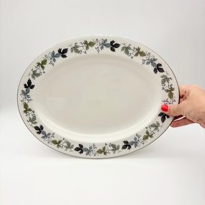 May include: An oval ceramic serving platter with a white center and a decorative border featuring a repeating pattern of dark green, light green, and grey leaves interspersed with small clusters of dark blue berries along the outer rim.