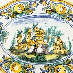 Pu&ograve; includere: Un piatto in ceramica bianca con un bordo giallo e disegni floreali blu e verdi. Il centro del piatto presenta una scena dipinta a mano di un piccolo villaggio con edifici e alberi.