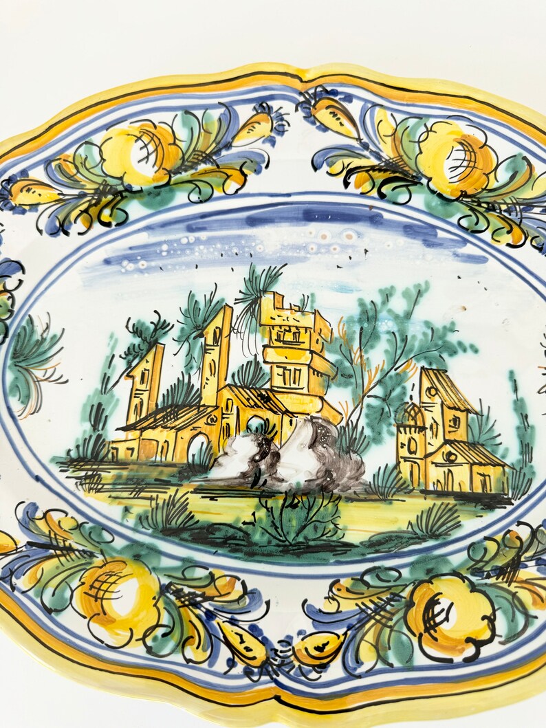 Pu&ograve; includere: Un piatto in ceramica bianca con un bordo giallo e disegni floreali blu e verdi. Il centro del piatto presenta una scena dipinta a mano di un piccolo villaggio con edifici e alberi.