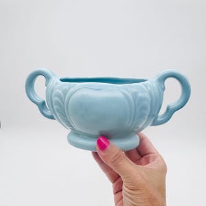 Pode incluir: Um vaso de cerâmica azul claro com duas alças curvas. O vaso apresenta um design em relevo e uma base arredondada. O objeto é segurado por uma pessoa com esmalte rosa.