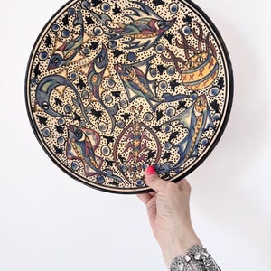 May include: A decorative ceramic plate with a black rim and a detailed fish design. The plate features colorful fish illustrations in blue, red, yellow, and green against a cream background. The plate is held up against a white background.