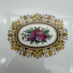 May include: White square plate with a floral design. The center features an oval with pink and purple roses, green leaves, and blue accents. The oval is framed by a brown border and surrounded by gold-colored decorative elements.