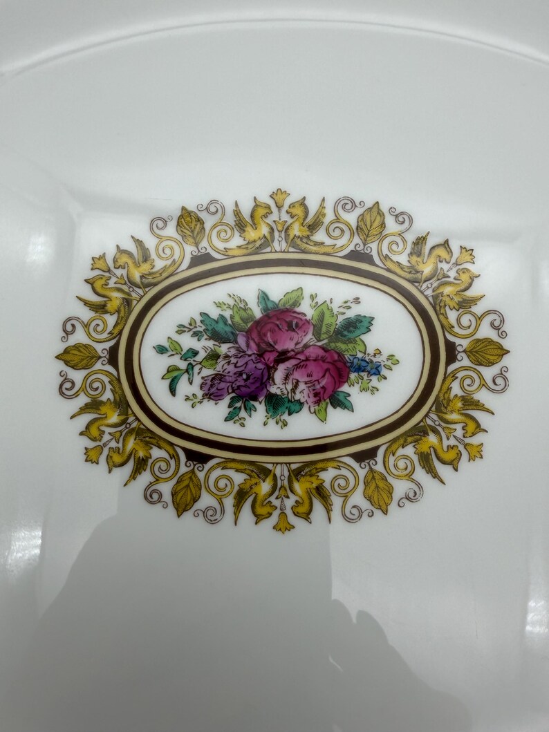 May include: White square plate with a floral design. The center features an oval with pink and purple roses, green leaves, and blue accents. The oval is framed by a brown border and surrounded by gold-colored decorative elements.