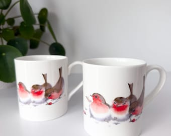 Könnte beinhalten: Zwei weiße Keramikbecher mit einer Aquarellillustration von drei kleinen Vögeln mit roten Brustfedern und braunen Flügeln. Die Vögel sind in einer Reihe auf einer weißen Fläche dargestellt, jeder Becher verfügt über einen geschwungenen Henkel.