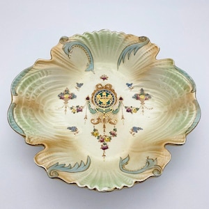 Könnte beinhalten: Eine antike, cremefarbene Porzellanschale mit gewelltem Rand. Die Schale ist mit einem grünen, goldenen und blauen Blumendesign und einem zentralen Wappen verziert.