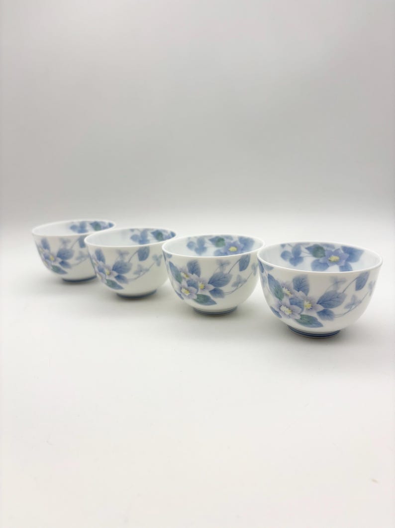 K&ouml;nnte beinhalten: Vier wei&szlig;e Keramik-Teetassen mit blauem Blumendesign. Die Tassen haben eine abgerundete Form und einen kleinen blauen Sockel. Das Blumenmuster umfasst blaue Bl&uuml;ten und gr&uuml;ne Bl&auml;tter. Die Tassen sind in einer Reihe angeordnet.