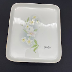 May include: White porcelain serving tray with a gold rim and a floral design featuring white flowers and green leaves. The tray is signed "Deline Style."