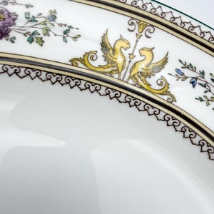 May include: Close-up of a white porcelain plate with a decorative border. The border features a repeating pattern of black and gold scrollwork, with yellow winged creatures and floral accents. The plate has a green rim.