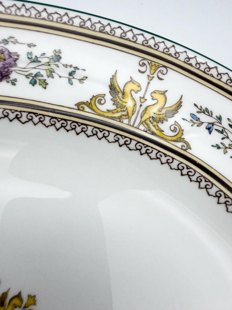 May include: Close-up of a white porcelain plate with a decorative border. The border features a repeating pattern of black and gold scrollwork, with yellow winged creatures and floral accents. The plate has a green rim.